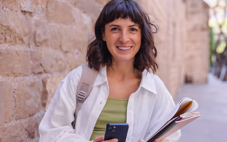 A smiling woman holding a smartphone in one hand and a notebook in the other, standing in front of a stone wall outdoors. She is wearing a white shirt over a green top and has a backpack strap on her shoulder.